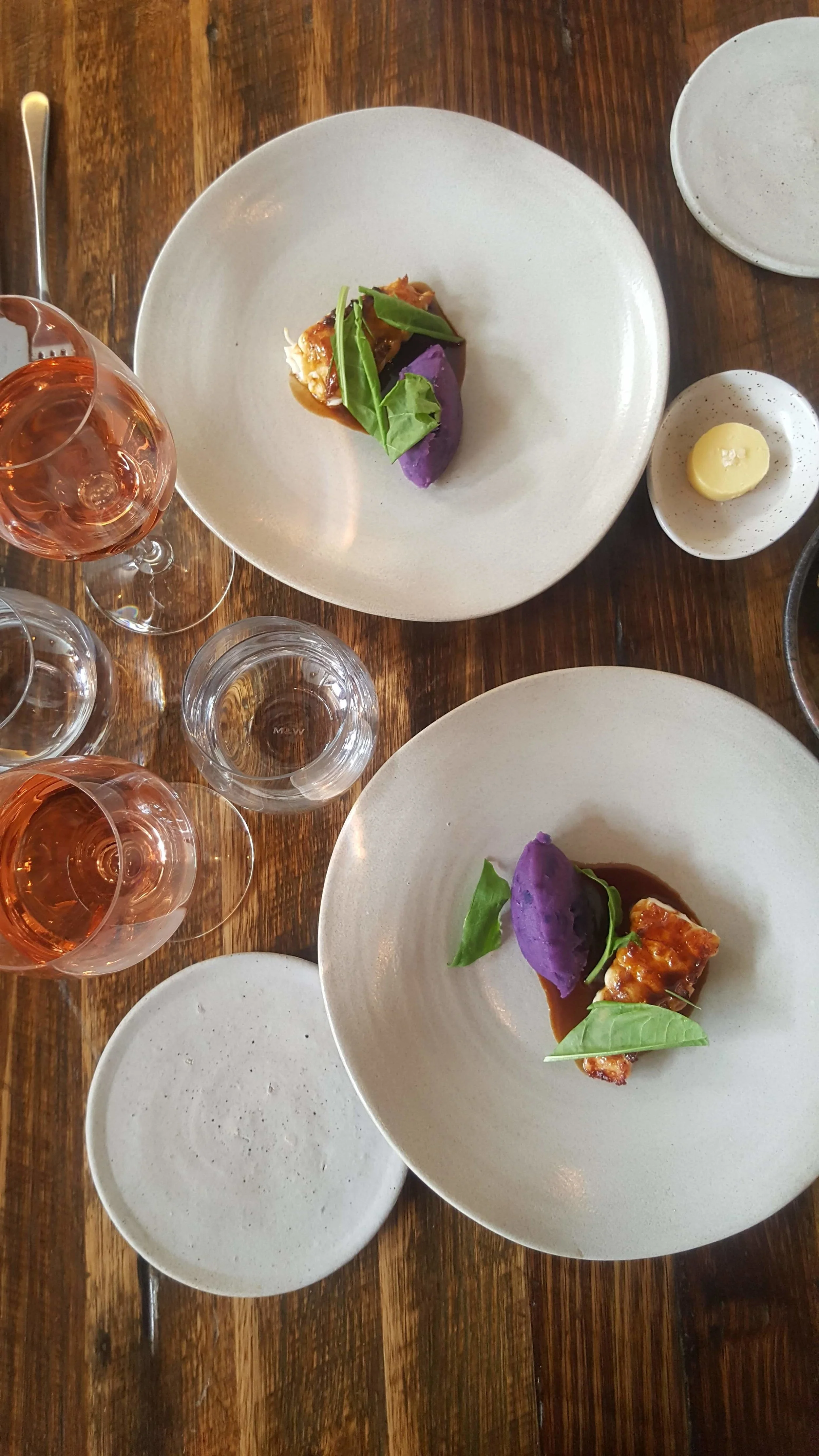 Two plated dishes with greens and sauce on a wooden table beside glasses, relaxed dining mood